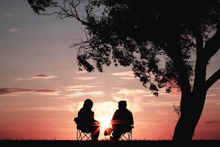 Image: two people in chairs sit beneath a tree as the sun rises